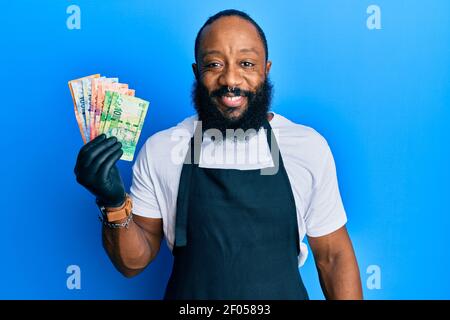 Junger afroamerikanischer Mann mit professioneller Schürze, der Südafrika hält rand Banknoten sehen positiv und glücklich stehen und lächeln mit Ein Confi Stockfoto