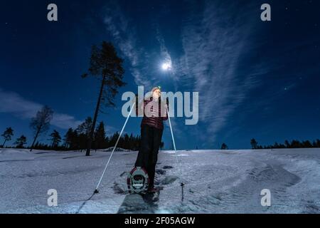 Schöne Frauen mittleren Alters kaukasischen in Magenta Dunstjacke und Schneeschuhe steht in der Nacht selten verschneiten Winterwald unter Vollmond Licht und sieht t Stockfoto