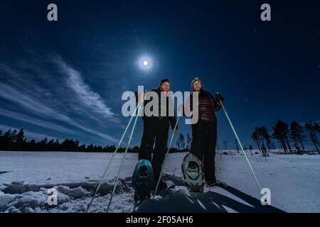 Schöne Männer und Frauen mittleren Alters in Schneeschuhen stehen in der Nacht selten verschneiten Winterwald unter Vollmondlicht. Nachtwanderung, Lappland, Umea, Schweden Stockfoto