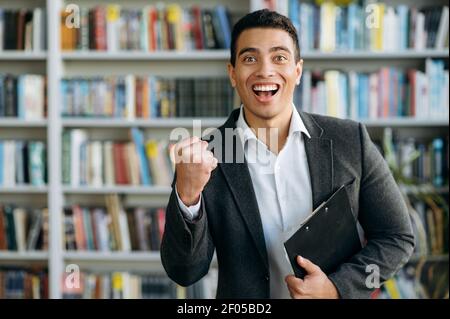 Überglücklich ist der junge Geschäftsmann über die Arbeitsförderung, schaut lächelnd auf die Kamera. Aufgeregt männlichen Unternehmer erstochen in modernen Büro, oder Student in einer Bibliothek Stockfoto