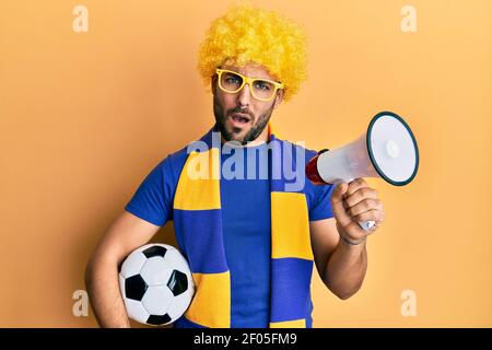 Junge hispanic Mann Fußball-Fan hält Fußball mit Megaphon im Schockgesicht, skeptisch und sarkastisch, überrascht mit offenem Mund Stockfoto
