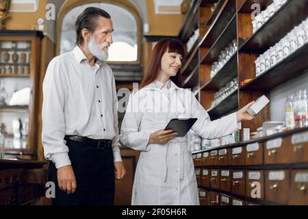 Junge attraktive kaukasische Apothekerin im Gespräch mit angenehmen älteren männlichen Kunden in alten vintage Apotheke, bietet gute Sicherheitsmedizin Stockfoto