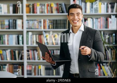 Fröhliche junge Erwachsene hispanic Freiberufler oder Student steht in modernen Büro oder Bibliothek, mit Laptop. Fröhlicher männlicher Angestellter in stilvollem Anzug posiert, schaut lächelnd auf die Kamera Stockfoto