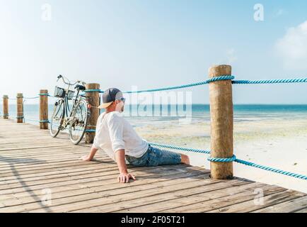 Portrait von entspannenden Mann in leichten Sommerkleidung gekleidet und Sonnenbrillen sitzen und genießen Sie Zeit und Blick auf den Strand auf Holz pier. Sorgloser Urlaub in t Stockfoto