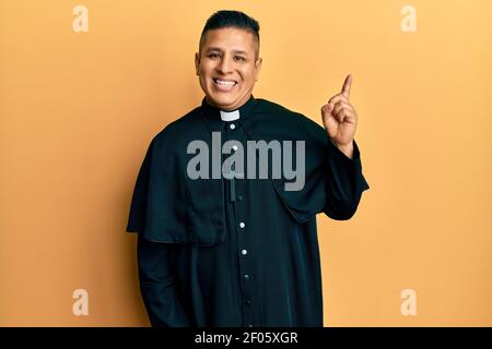 Junger lateinischer Priester, der auf gelbem Hintergrund mit einem großen Lächeln auf dem Gesicht steht und mit dem Handfinger zur Seite zeigt und auf die Kamera schaut. Stockfoto