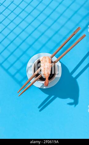 Nigiri Garnele in der Nähe von Sojasauce, mit Essstäbchen auf blauem Hintergrund. Hartes Licht und Schatten. Traditionelle japanische Küche. Speisekarte im Restaurant, Sushi-Bar Stockfoto