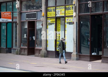 Ein Mann geht an einer Reihe geschlossener Geschäfte in der Queen Street vorbei, die während der Sperrzeit des Coronavirus in Cardiff, Wales, Großbritannien, ein Gesicht tragen. Stockfoto