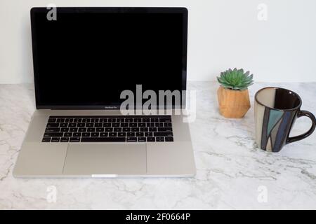Mac Laptop sitzt auf einer Marmoroberfläche mit schwarzem Bildschirm, Tasse und kleine Pflanze daneben. Stockfoto