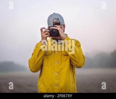 Junger männlicher Fotograf in gelber Jeansjacke, der fotografiert Fotokamera im Stehen auf neblig verschwommenem Naturhintergrund Stockfoto