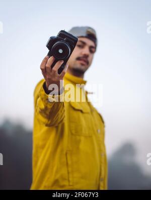Side view content junger männlicher Fotograf in gelber Jeanjacke Fotografieren mit der Fotokamera und betrachten der Kamera während Stehen auf nebligen verschwommen na Stockfoto