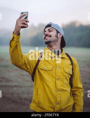 Lächelnder junger männlicher Backpacker in gelber Jeansjacke, der Selfie nimmt Auf dem Smartphone im Stehen auf nebliger Wiese in der Natur Stockfoto