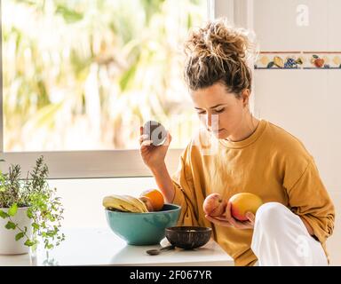 Nachdenkliche junge Frau mit lockigem Haar in legerer Kleidung sitzt am Tisch und wählt frisches Obst für ein gesundes Frühstück zu Hause bei Tageslicht Stockfoto