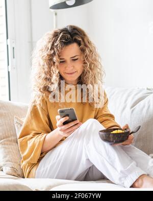 Lächelnde junge Frau in lockigem Haar in legerer Kleidung auf einem bequemen Sofa sitzend und Messaging auf dem Smartphone, während sie im hellen Raum gesunde Lebensmittel isst Stockfoto