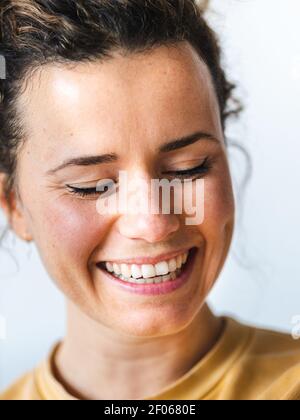Positive Frau in legerer Kleidung lächelnd mit geschlossenen Augen gegen verschwommenes Licht Wand in der Wohnung Stockfoto