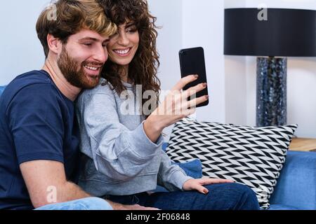 Junger bärtiger Mann, der einer lächelnden Frau in legerer Kleidung nahe kommt und Selfie auf dem Mobiltelefon nimmt Stockfoto