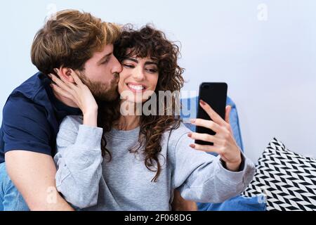 Junger bärtiger Mann küsst lächelnde Frau in legerer Kleidung und nimmt Selfie auf dem Handy Stockfoto