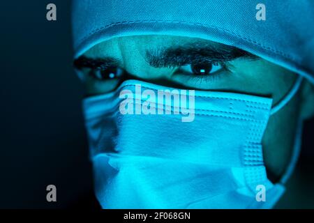 Nahaufnahme des männlichen Chirurgen in der medizinischen Maske wegschauen in Dunkles Zimmer mit blauem Neonlicht Stockfoto