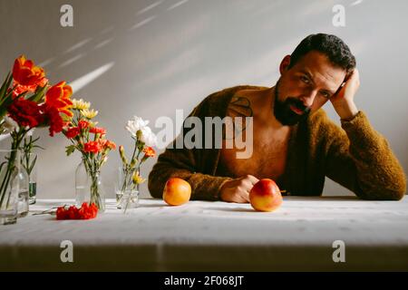 Bärtig nachdenklich männlich lehnte auf der Hand sitzen am Tisch mit Reife Äpfel und Blumen in Gläsern Stockfoto