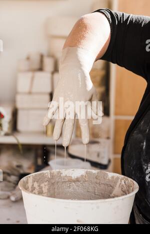 Ernte anonyme Handwerker in schmutzigen schwarzen Kleidern, die neben dem Tisch stehen und schmutzige Hand aus dem Eimer ziehen, der mit Lehm gefüllt ist, im Studio bei Tageslicht Stockfoto