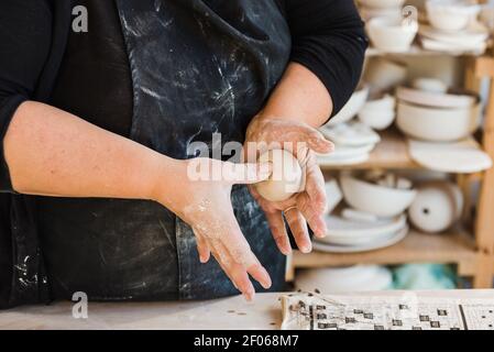 Crop anonyme Meister in schwarz schmutzige Schürze in der Werkstatt stehen Und das Formstück Ton in den Händen neben dem Tisch mit Werkzeuge zum Basteln Stockfoto