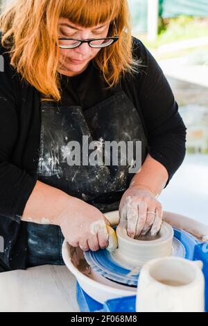 Fokussierte Frau, die mit einem Wurfrad neben dem Tisch steht, während sie in der Werkstatt mit Lehm modelliert Stockfoto