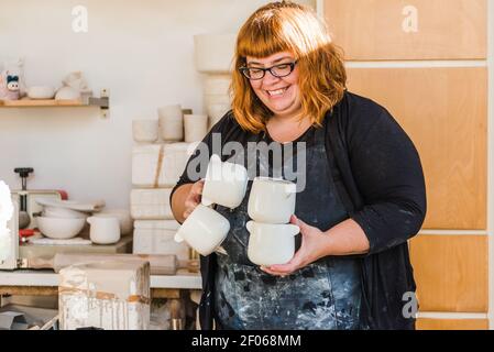 Lächelnde Erwachsene Kunsthandwerkerin in schmutziger Schürze und schwarzer Kleidung Stehen im Lichtstudio und tragen handgemachte Keramik Töpfe in der Nähe Clayware Stockfoto