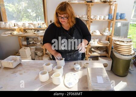 Adult female artisan in dirty apron and black clothes standing in light studio with handmade ceramic pots near clayware Stockfoto