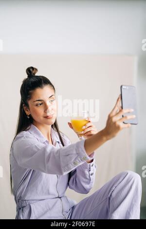 Junge freundliche Frau mit einem Glas Orangensaft, der sich selbst nimmt Porträt auf Handy im Haus Stockfoto