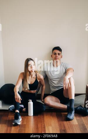 Ganzkörper aus Mann und Frau in sportlicher Kleidung mit gekreuzten Beinen auf Parkett in der Nähe der Wand mit einer Flasche Wasser nach dem Training sitzen Stockfoto