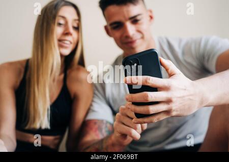 Content Mann und Frau tragen Sportkleidung Surfen Handy, während auf dem Boden gekreuzt Beinen in der Nähe der Wand mit einer Flasche Wasser sitzen Stockfoto