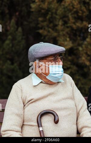 Porträt eines älteren Mannes mit Gesichtsmaske und Baskenmütze suchen Unterwegs auf einer Parkbank sitzen Stockfoto