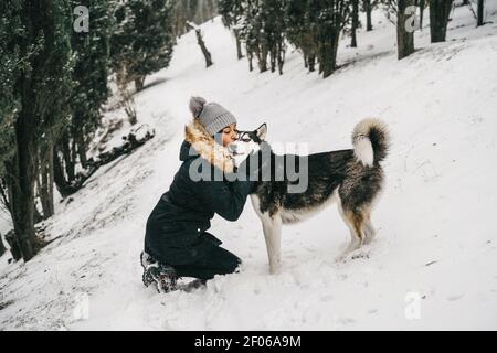 Lächelnd junge ethnische Dame trägt Oberbekleidung Umarmung und küssen niedlich husky Hund beim Hocken in verschneiten Wäldern in der Nähe von grünen Fichten Im Winter Tag Stockfoto