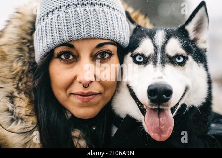 Junge ethnische Dame trägt Oberbekleidung Umarmung niedlich Husky Hund während Stehen in verschneiten Wäldern in der Nähe von grünen Fichten im Winter Tag Stockfoto