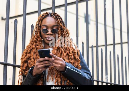 Von unten afroamerikanische Frau in stilvollem Outfit angelehnt Zaun und Surfen auf dem Smartphone auf der Stadtstraße Stockfoto