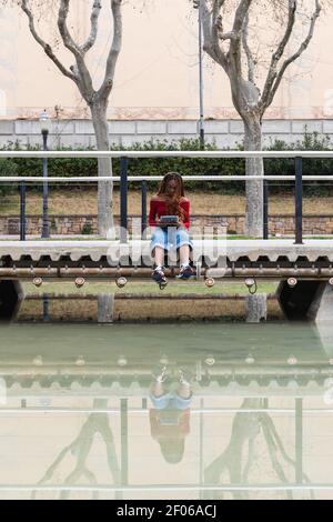 Afroamerikanische Frau, die auf einer Fußgängerbrücke in der Nähe des Sees in der Stadt sitzt Und Surfen im Internet auf Tablet Stockfoto