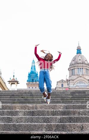 Sorglos lächelnde afroamerikanische Frau, die Musik in Kopfhörern hört Und springen über Treppen in der Stadt, während Sie die Kamera betrachten Und zeigt Rock ge Stockfoto