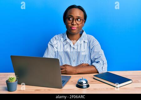 Junge afrikanerin, die an der Hotelrezeption mit einem Laptop arbeitet, lächelt und schaut zur Seite und starrt wegdenkend. Stockfoto