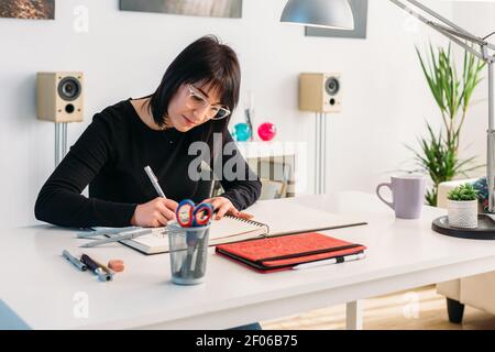 Fokussierte talentierte Künstlerin Zeichnung in Notizblock während sitzen Tisch im Wohnzimmer zu Hause Stockfoto