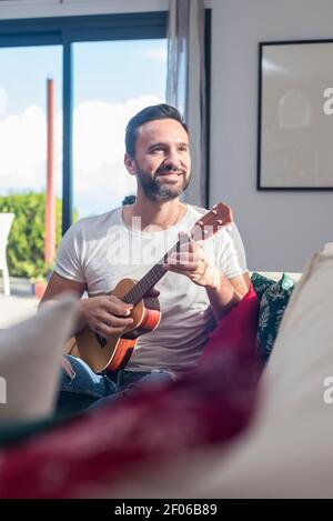 Positiver bärtiger ethnischer Gitarrist, der lächelt, während er Ukulele sitzend spielt Auf Sofa in moderner Wohnung Stockfoto