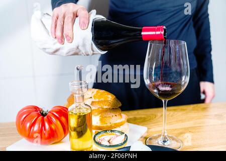 Anonyme Person serviert Rotwein in einem typischen spanischen oder katalanischen Essen. Brot mit Tomaten, iberischem Schinken, Ziegenkäse, Öl und Knoblauch. Ein Abendessen vorbereiten Stockfoto