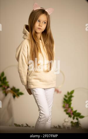 Nachdenkliche Mädchen 8-9 Jahre alt in einem beigen Sweatshirt und weißen Hosen, hält ihre Hände hinter einem Bett auf einem cremefarbenen Hintergrund Stockfoto