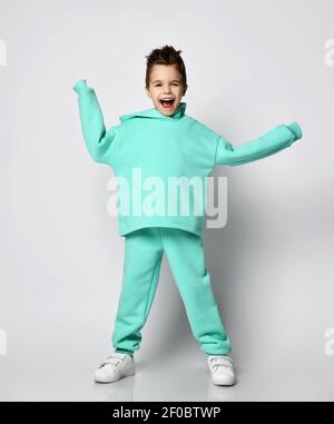 Happy Screaming Kid Boy in modernen grünen, mintfarbenen Hoodie, Hosen und weißen Sneakers steht mit Händen in den Ärmeln Stockfoto