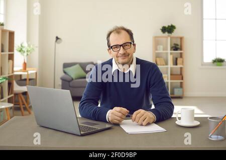 Glücklicher Mann mittleren Alters in einer Brille lächelt auf die Kamera sitzen Am Schreibtisch in seinem Heimbüro Stockfoto
