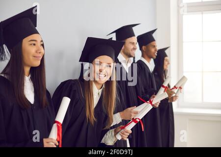 Frau mit einem Diplom in der Hand steht in einer Reihe in der Nähe seiner multiethnischen Klassenkameraden Freunde in der Nähe der Wand. Stockfoto