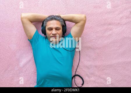 Der 50-jährige Mann hört Musik mit Kopfhörern zu Hause, auf dem Bett liegend. Entspannend, genießend. Stockfoto