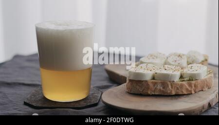 Weizenbier in einem Glas neben Ciabatta-Sandwiches mit Mozzarella und Pesto auf Leinentuch gießen, breites Foto Stockfoto