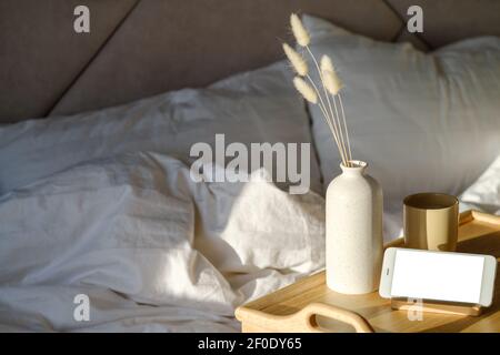 Frühstück im Bett Konzept. Tablett mit getrockneten Blumen in Vase, Tasse Kaffee, Smartphone Mockup. Weiß gestreifte Decke und Kissen auf dem Hintergrund. Hygge, Scan Stockfoto