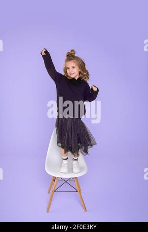 Schöne lustige Mädchen 6-7 Jahre alt tanzt auf Stuhl im Studio auf violettem Hintergrund. Konzept der Freude des Sieges, Überraschung. Foto in voller Länge Stockfoto