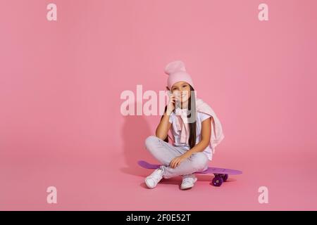 Kleines Mädchen sitzt auf Skateboard und mit Smartphone über Rosa Hintergrund Stockfoto