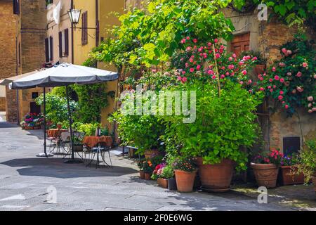 Wunderbares, süßes kleines Straßencafé mit Tischen und Stühlen in der alten gepflasterten Straße. Zierpflanzen und verzierte Wände mit bunten Blumen in Pienza Stockfoto